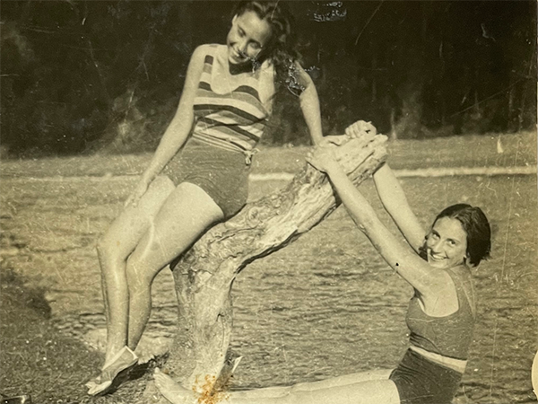 An old black-and-white photo of two women with a tree stem hanging over a lake. One woman is reclining on it while the other is hanging off it, and both are smiling.